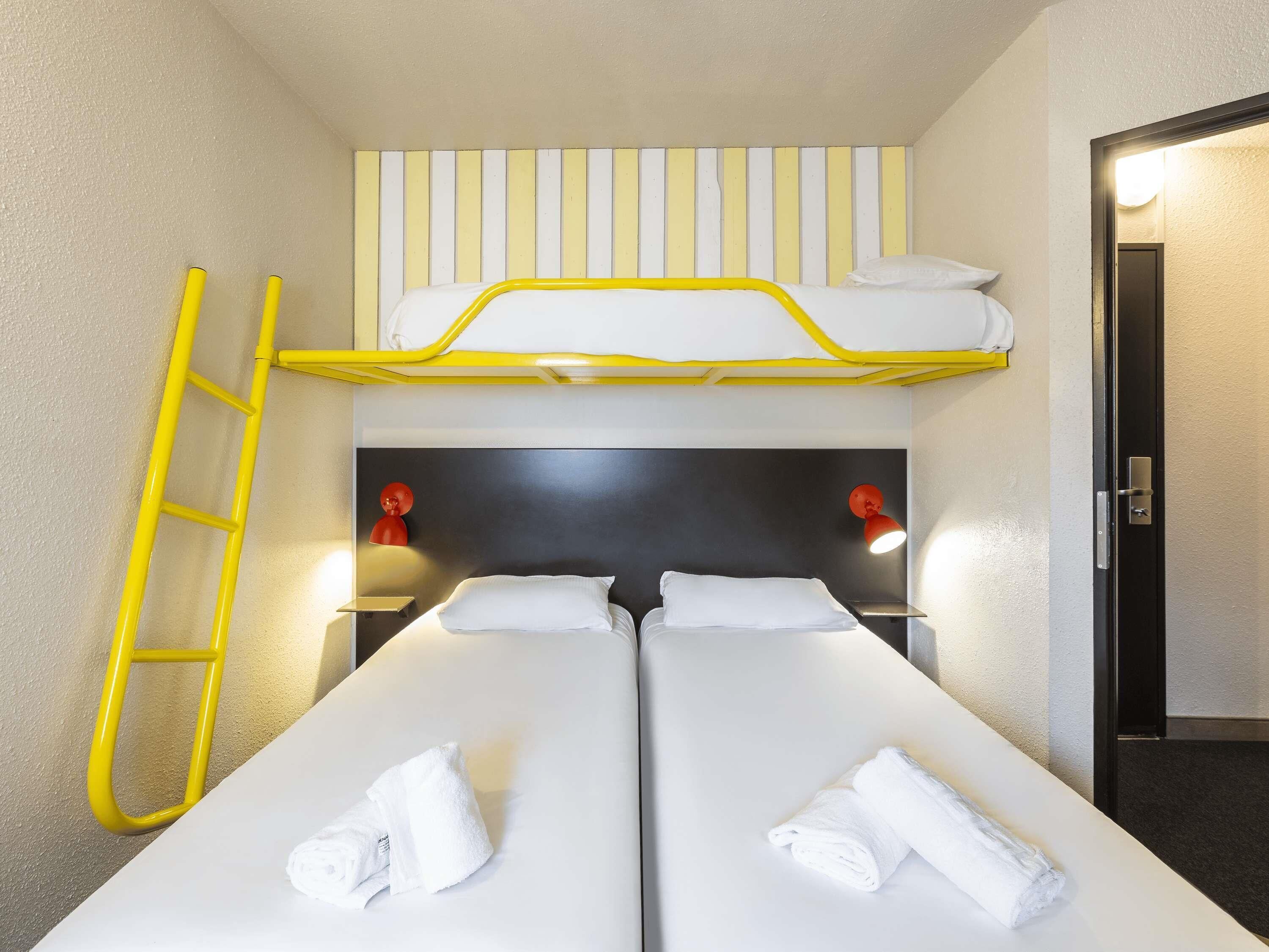 Una habitación con dos camas, una escalera amarilla, dos lámparas rojas en la pared y una puerta.
