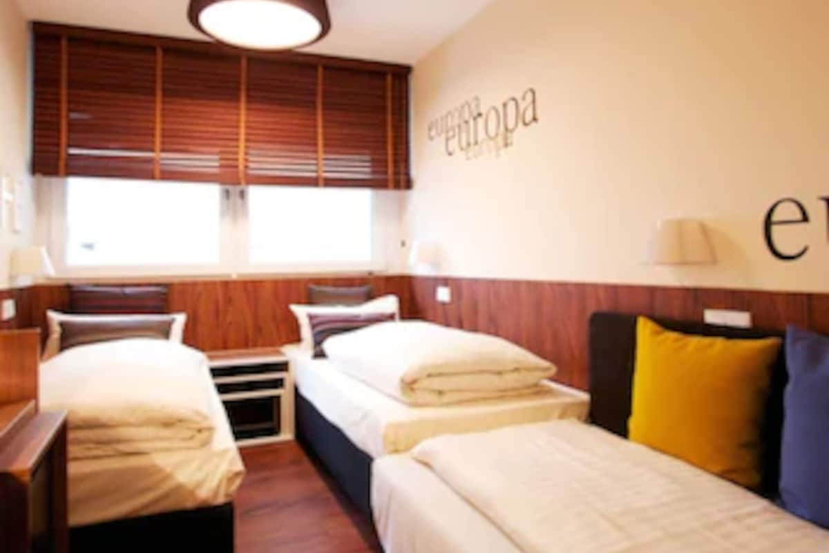 Hotellihuone, jossa on kaksi sänkyä, puulattia, kattovalaisin ja ikkuna, jossa on sälekaihtimet.
