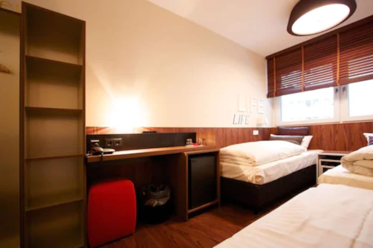 Hotellihuone, jossa on työpöytä, kaksi sänkyä, pieni punainen tuoli ja ikkuna, jossa on sälekaihtimet.