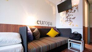 Une chambre d’hôtel moderne équipée d’un canapé-lit, d’une petite table de chevet, d’un téléviseur fixé au mur et d’un élément décoratif mural.