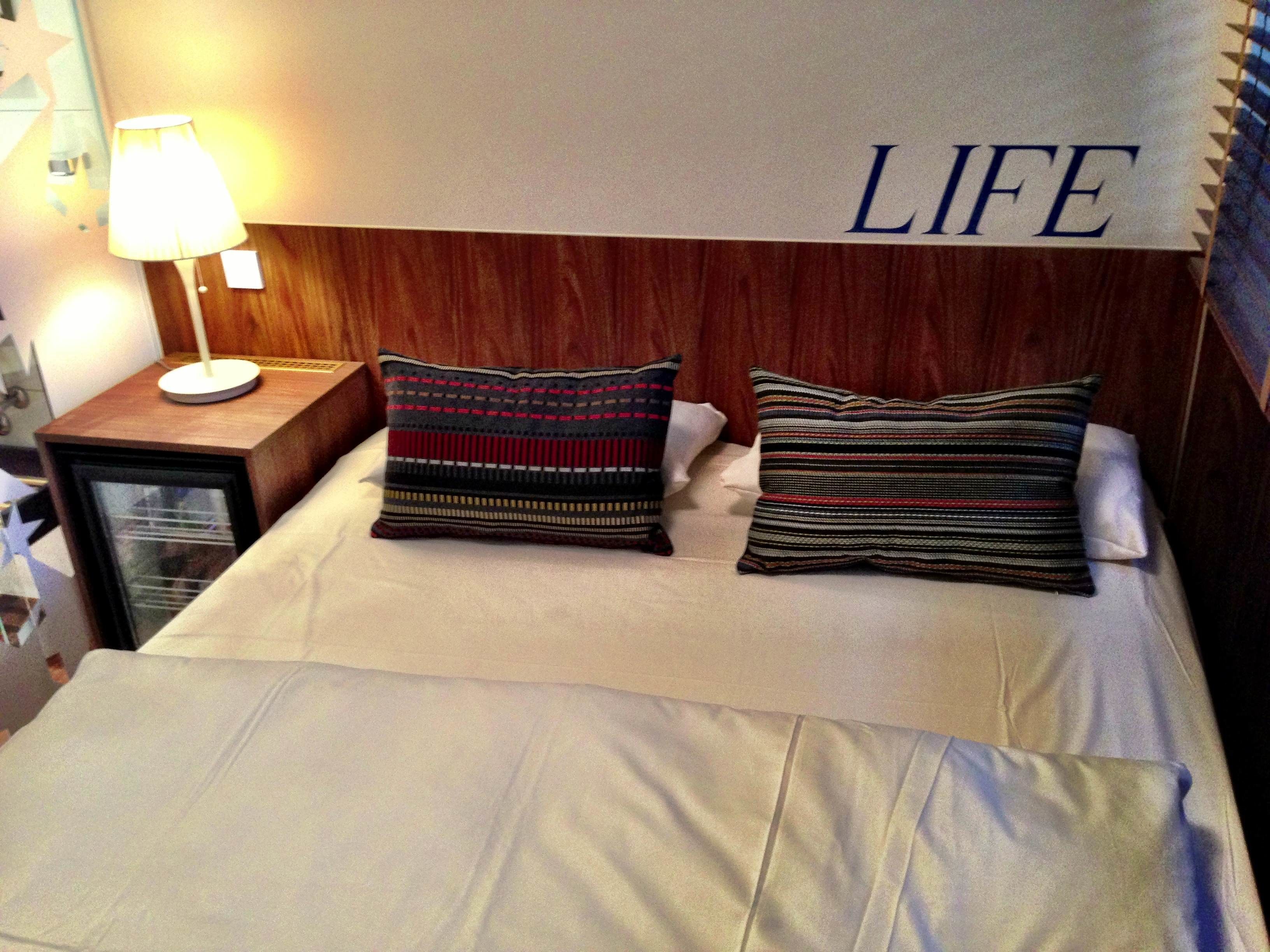 Ein Bett mit zwei Kissen, ein Nachttisch mit Lampe und ein Schild an der Wand mit der Aufschrift 'LIFE'.