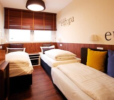 Habitación de hotel con dos camas, suelo de madera, ventana con persianas y una lámpara fijada a la pared.