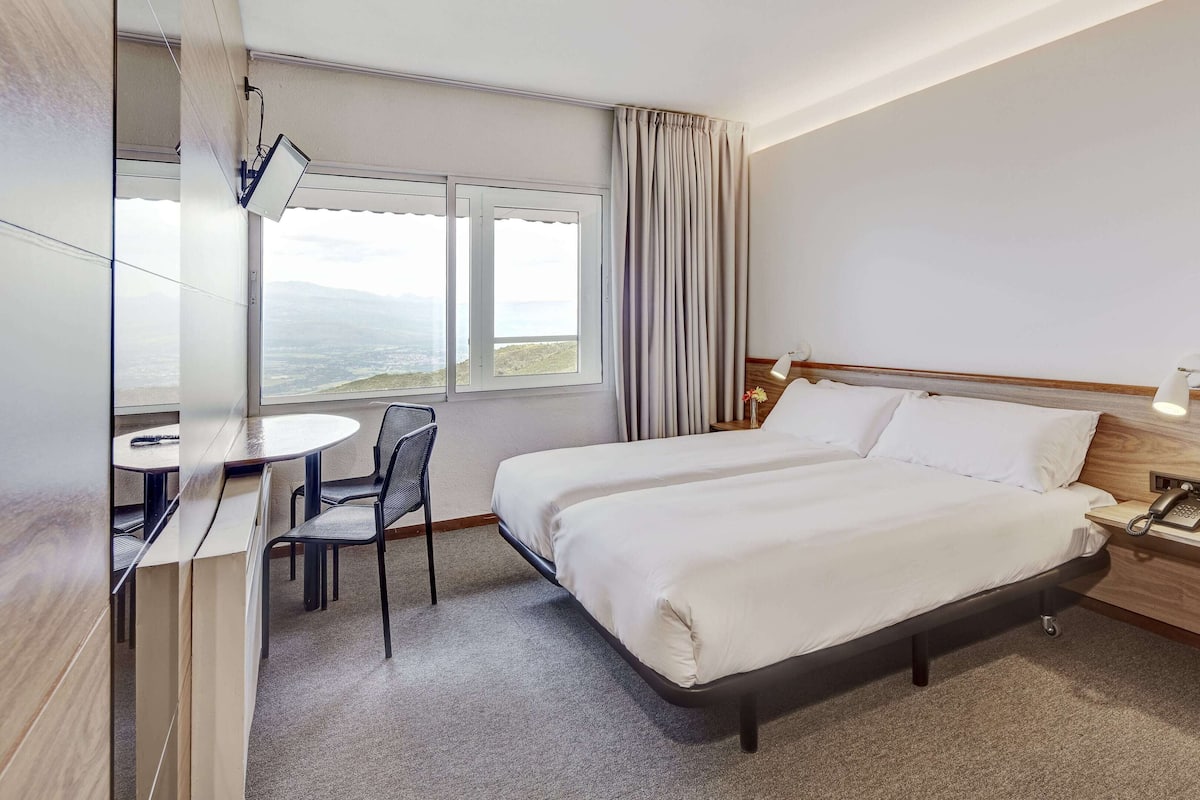 A hotel room with a bed, a desk, and a chair. There is a window with curtains, a nightstand with a lamp, and a phone.