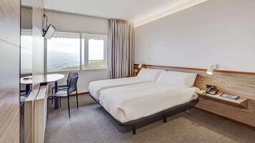 Habitación de hotel con cama, escritorio y silla. Hay una ventana con cortinas, una mesita de noche con lámpara y un teléfono.
