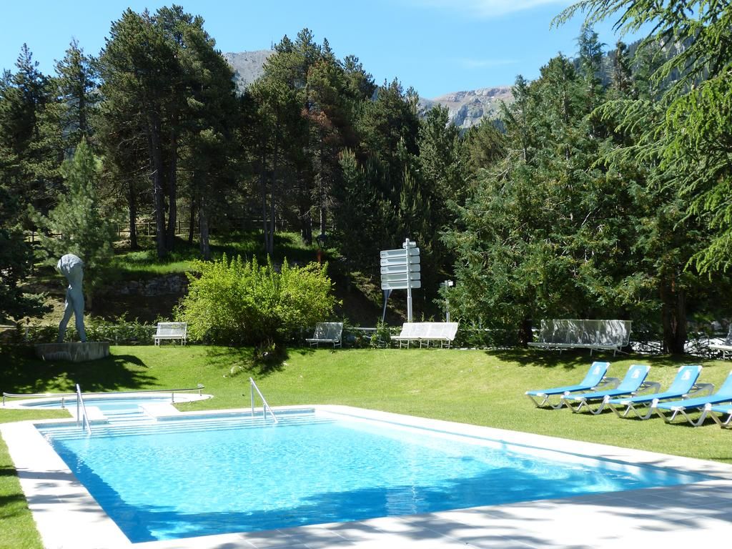 Una piscina con agua cristalina azul, rodeada de césped verde y árboles. Se ven sillas de descanso y una estatua al fondo.