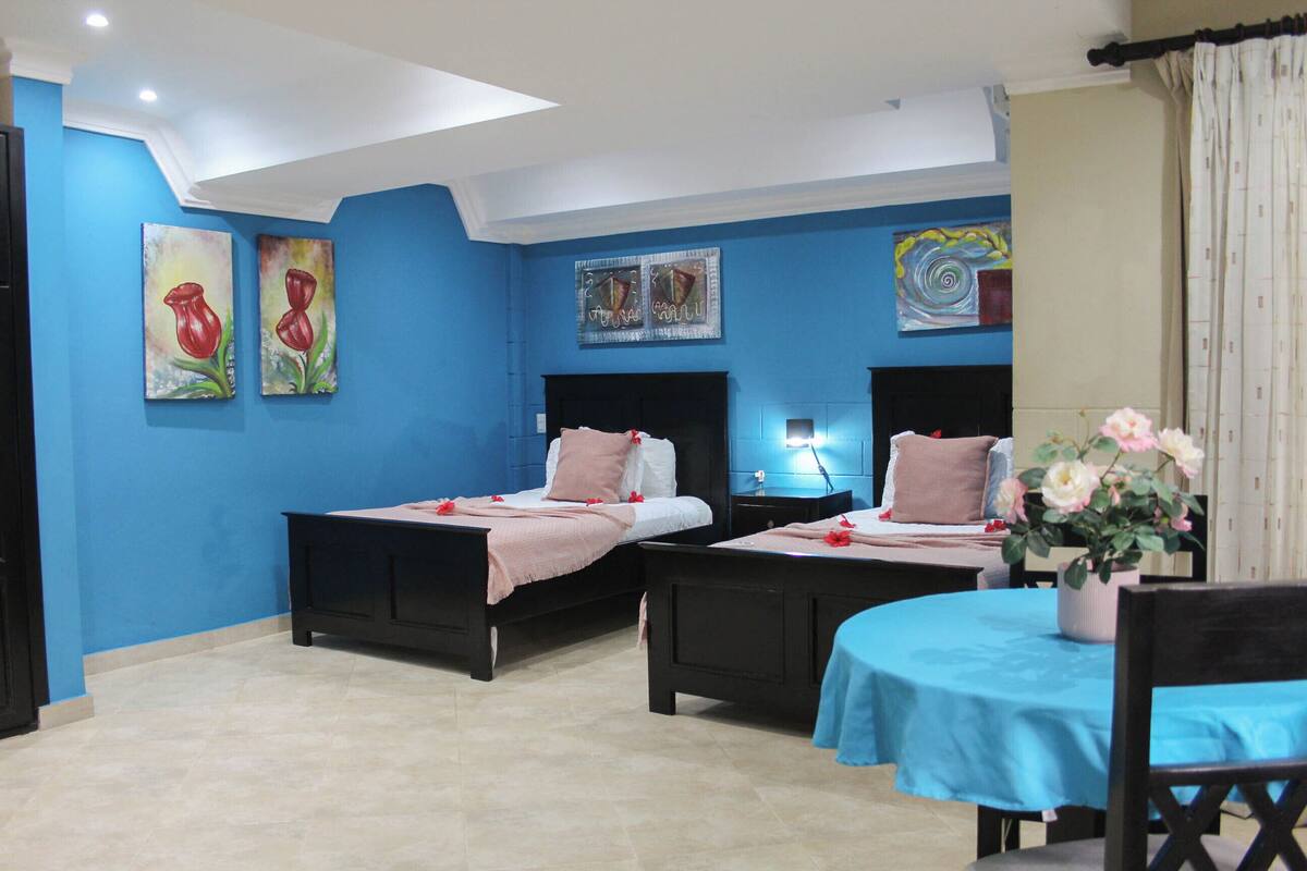 A hotel room with two beds, a small table, and a chair. There are paintings on the walls and a potted plant on the table.