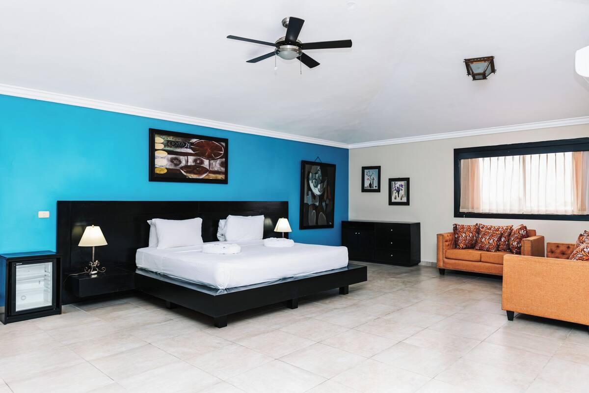 A bedroom with a bed, a ceiling fan, a nightstand, a lamp, a sofa, and a window with curtains.