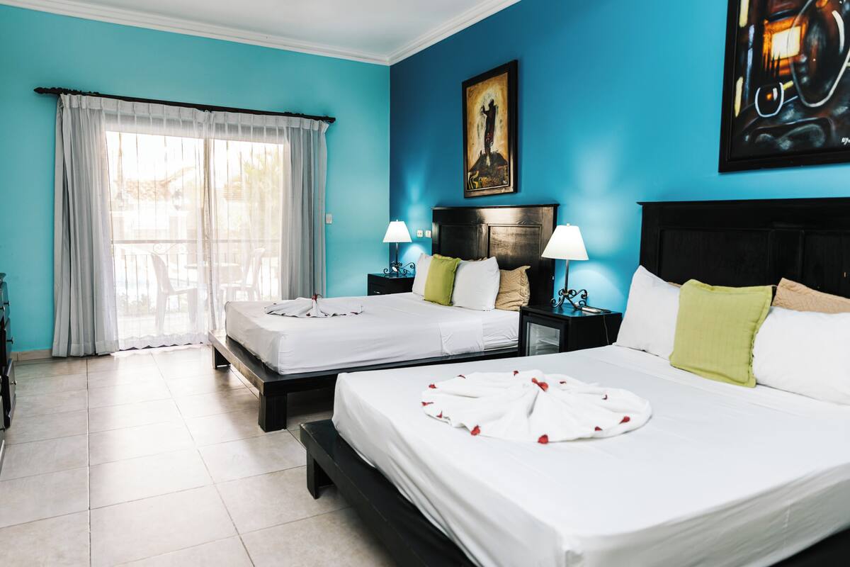 A hotel room with two beds, a large window with curtains, a painting on the wall, and a nightstand with a lamp.