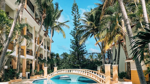 A pool area with a bridge, surrounded by palm trees and a multi-story building. - The MT Hotel (Punta Cana)