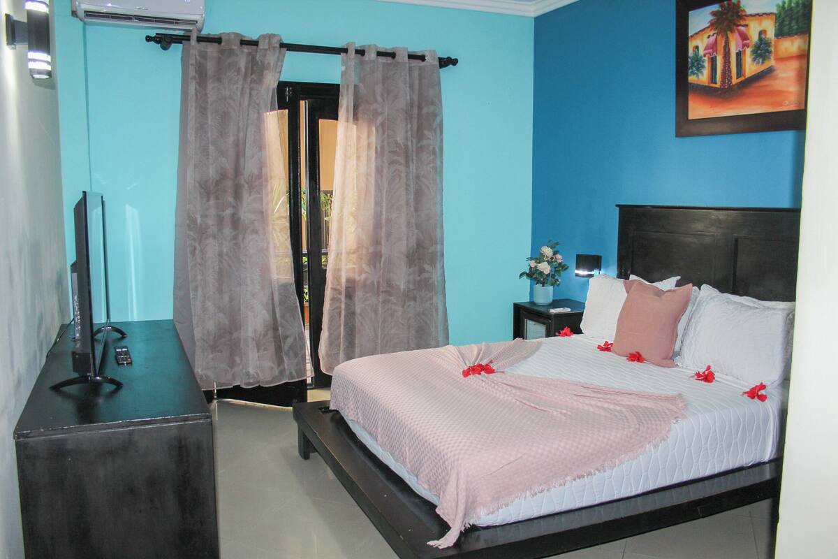 A bedroom with a bed, a nightstand, a TV, and a window with curtains.