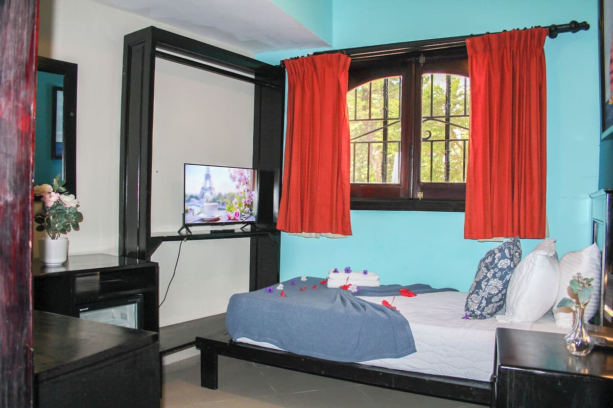 A bedroom with a bed, a television, a window with curtains, a dresser, and a vase with flowers.
