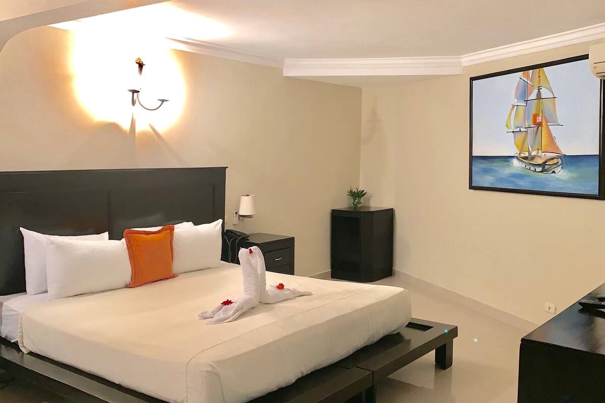 A modern hotel room with a neatly made bed, a bedside table with a lamp, a small plant, and a painting of a sailboat on the wall.
