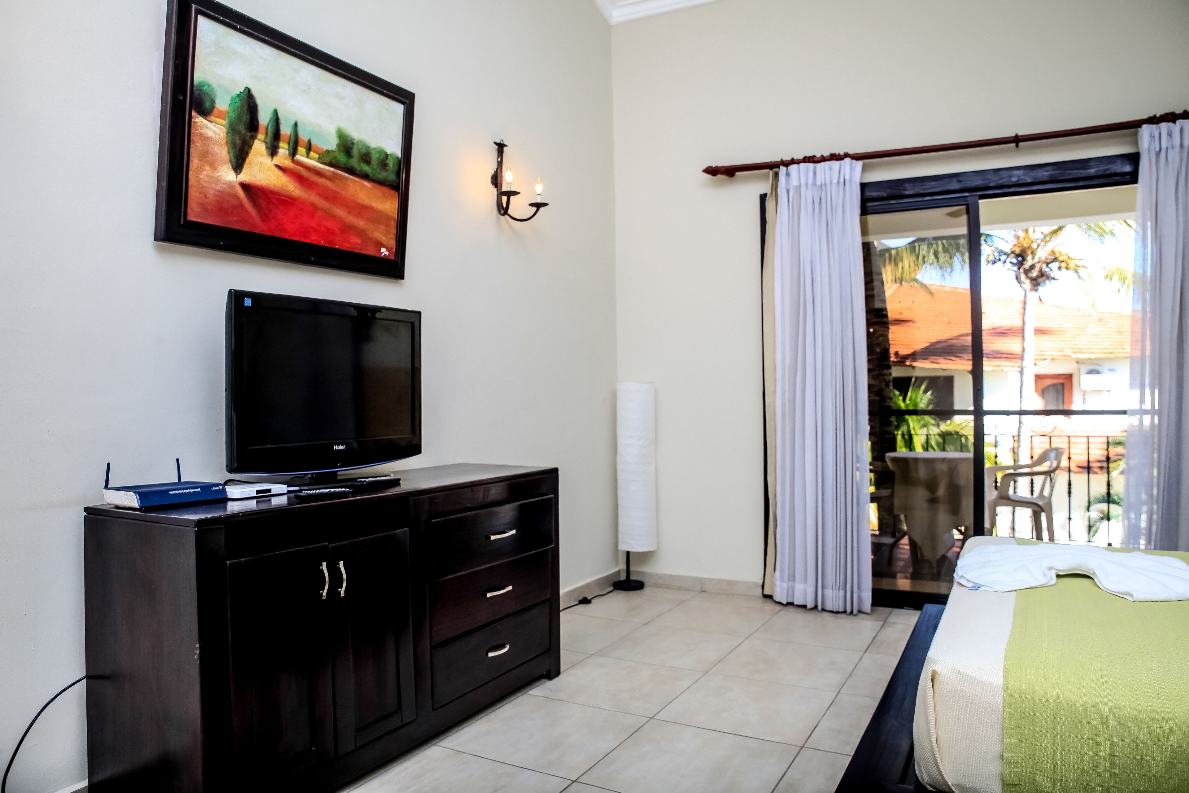 Habitación de hotel con cama, televisor sobre un mueble de madera oscura, un cuadro en la pared y vista al exterior a través de una puerta corrediza de vidrio.