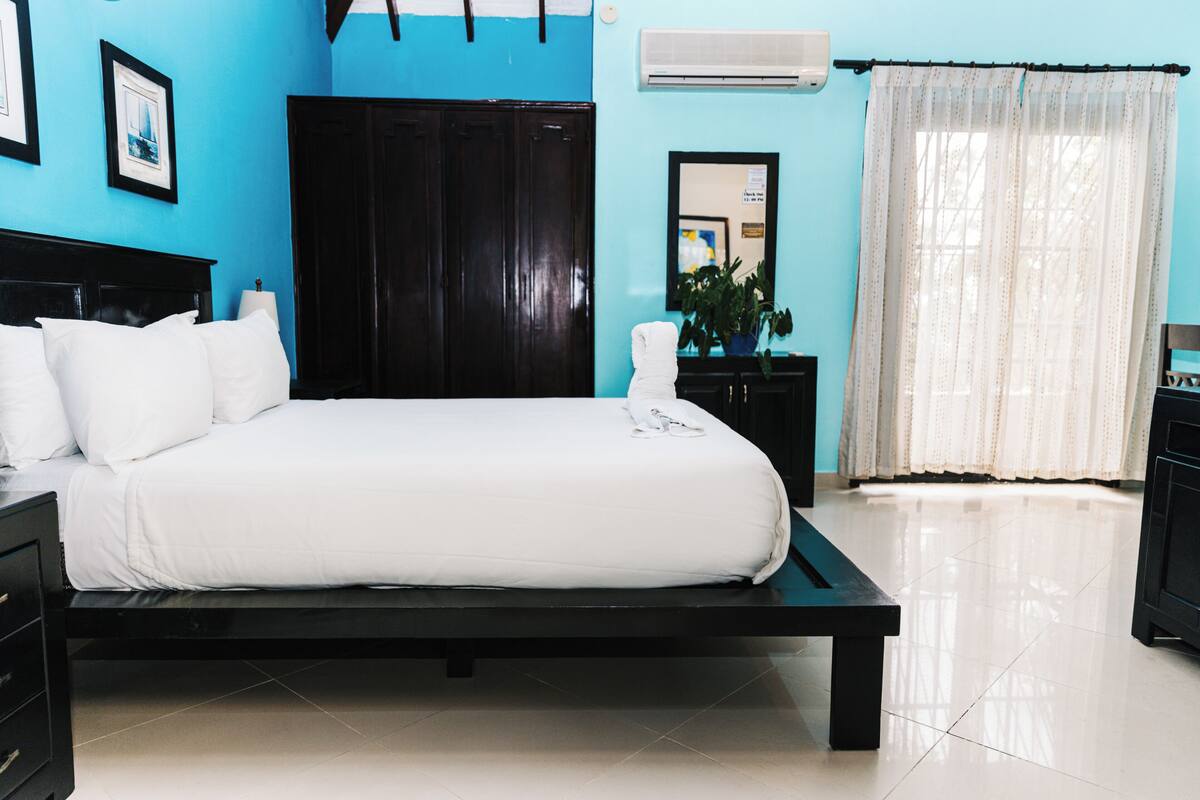 A neatly made bed with white bedding, a dark wooden headboard, a black nightstand, a potted plant, and a window with sheer curtains.