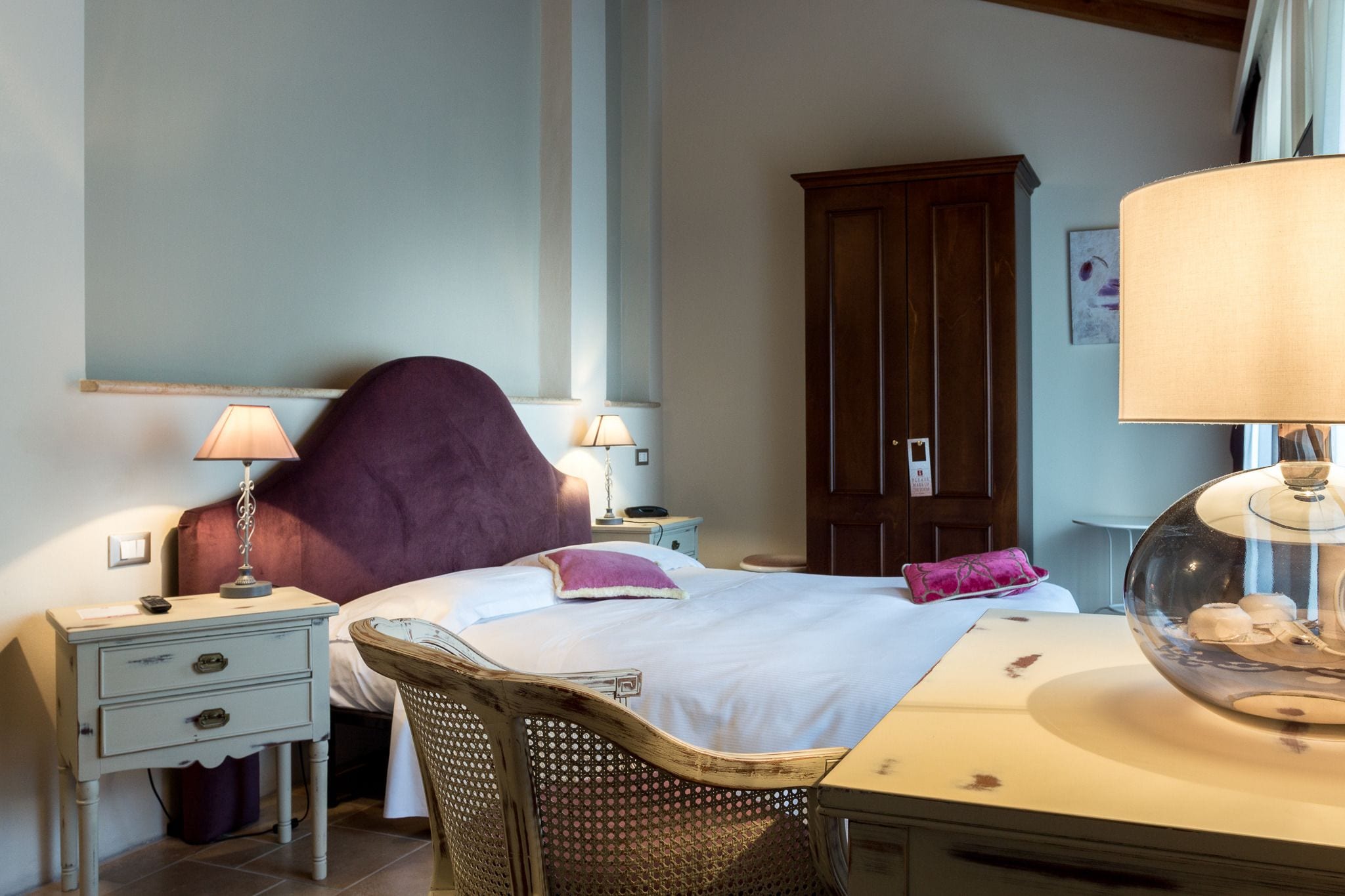 A bedroom with a bed, two bedside tables with lamps, a wooden wardrobe, and a wicker chair.