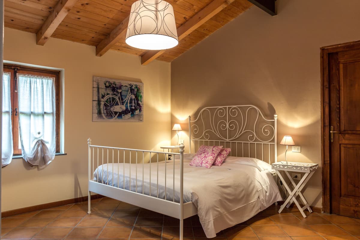 Een slaapkamer met een bed, nachtlampjes, een houten plafond, een raam met gordijnen en een deur.
