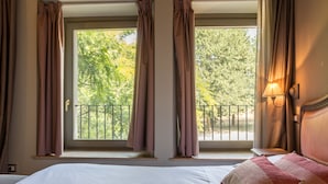 Une chambre d’hôtel avec un lit, deux fenêtres avec des rideaux, une vue sur les arbres et une table de chevet avec une lampe.