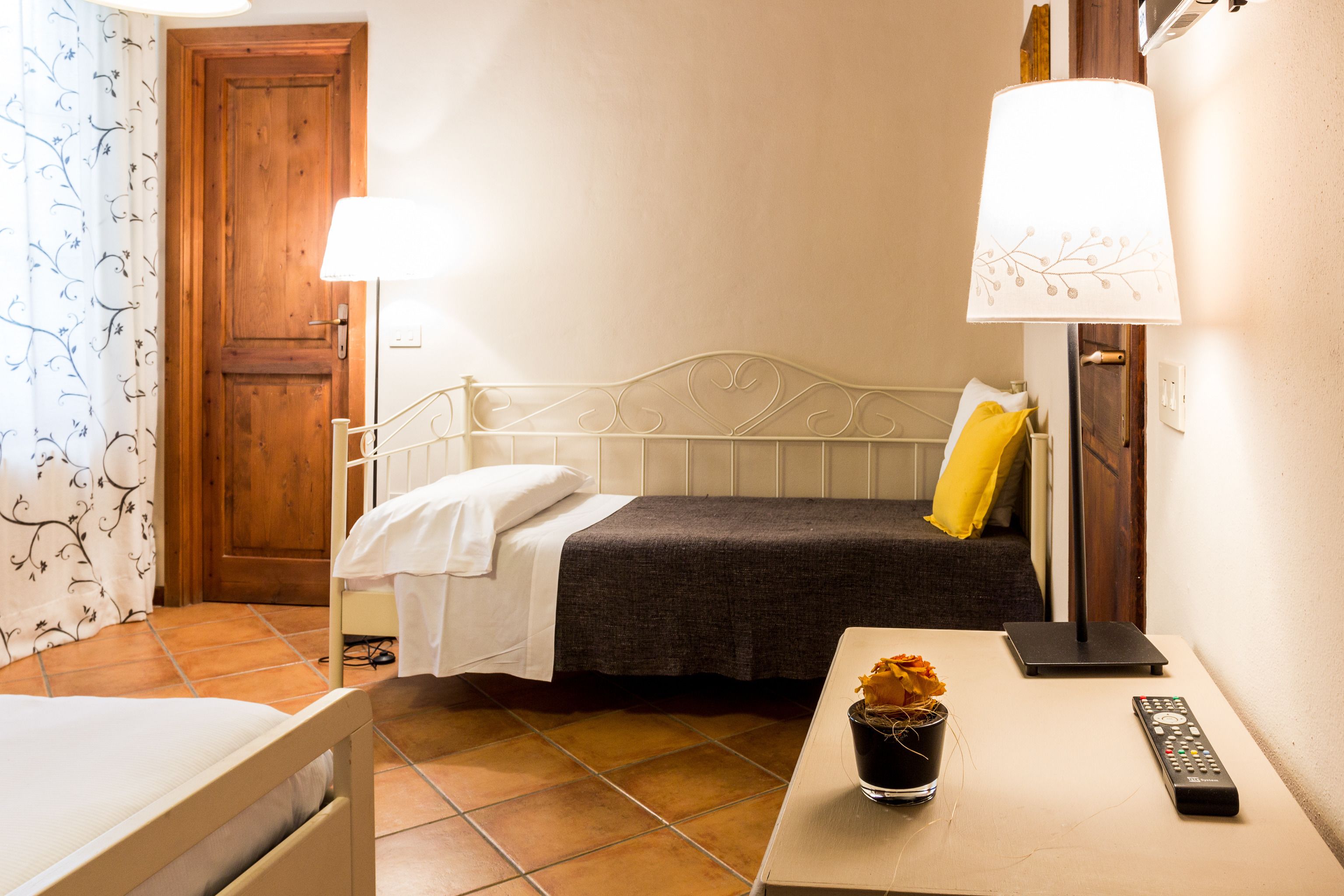 A hotel room with a bed, a bedside table with a remote control, a lamp, and a potted plant.
