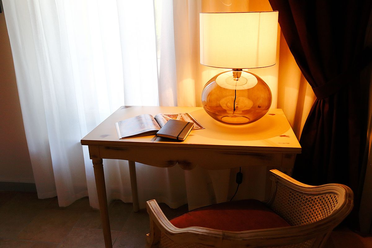 A hotel room with a wooden table, a lamp, a chair, and a window with curtains.