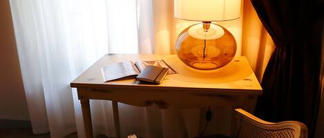 A hotel room with a wooden table, a lamp, a chair, and a window with curtains.