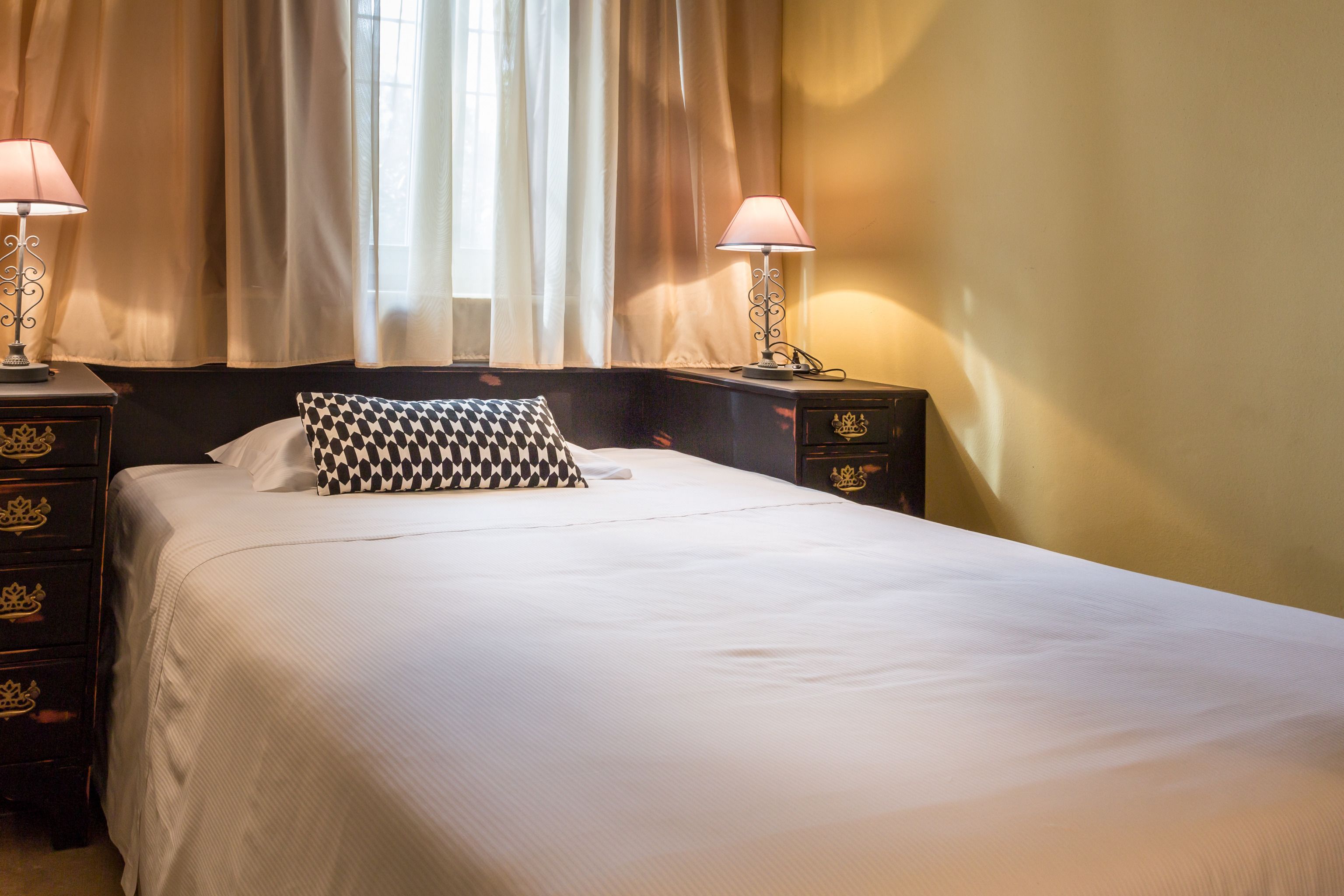A neatly made bed with a checkered pillow, two bedside lamps, and a nightstand with drawers.