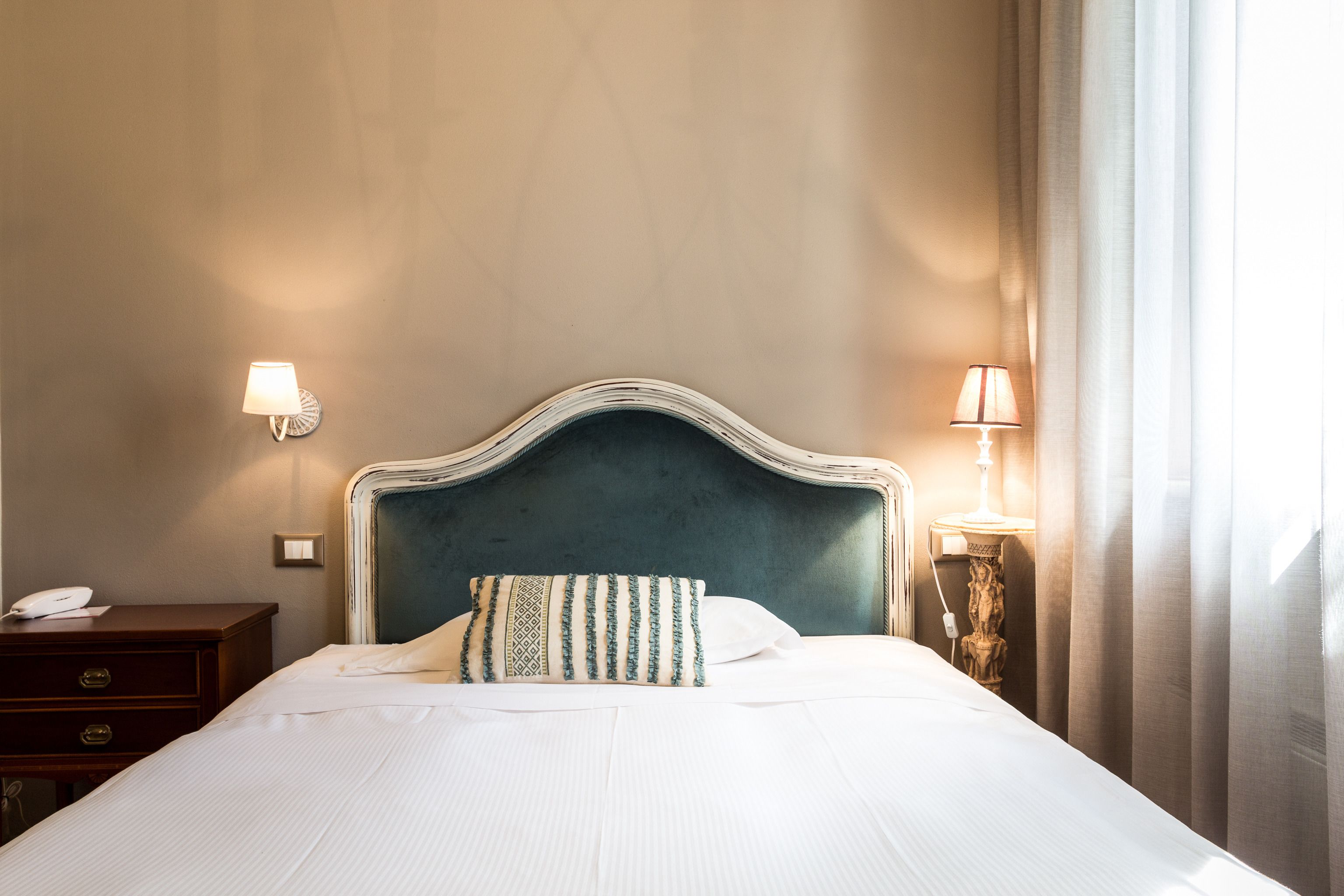 A neatly made bed with a patterned pillow, a bedside table with a lamp, and a wall-mounted light fixture.