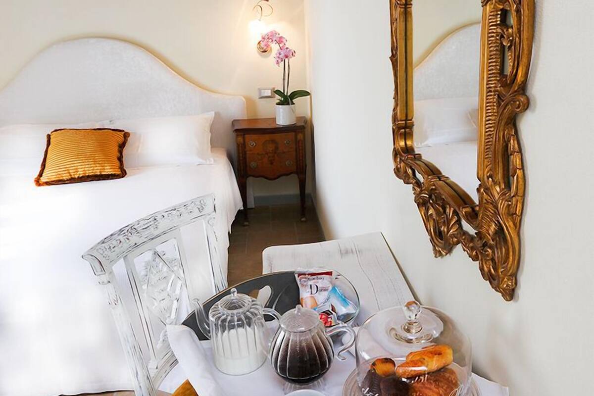 Hotelkamer met een bed, nachtkastje met lamp, dienblad met thee, koffie en gebak, en een grote, versierde spiegel aan de muur.