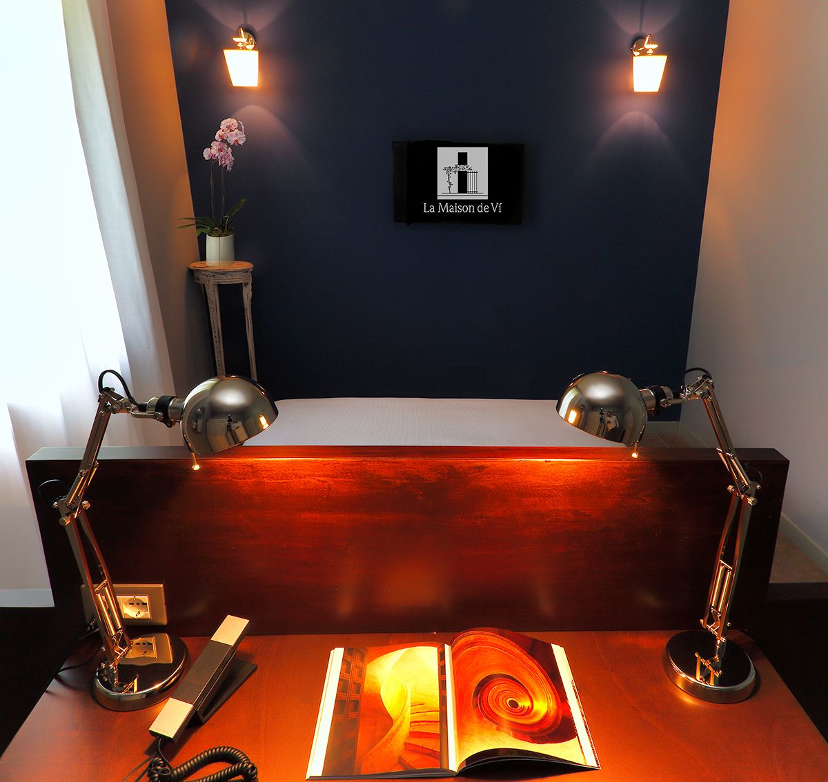 A room with a desk, two desk lamps, a framed sign on the wall, and a potted plant.