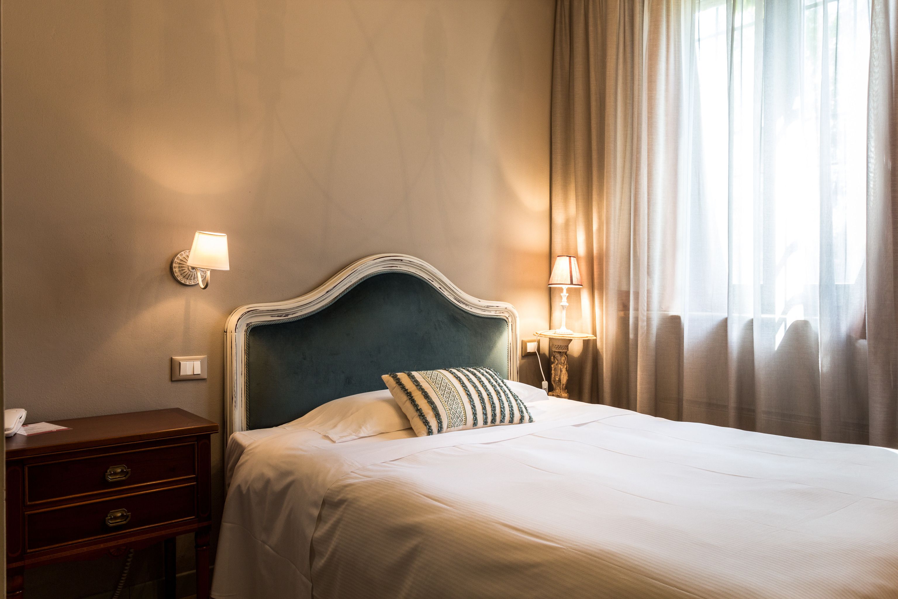 Una cama bien tendida con cabecera, una mesita de noche con lámpara y un aplique de pared.