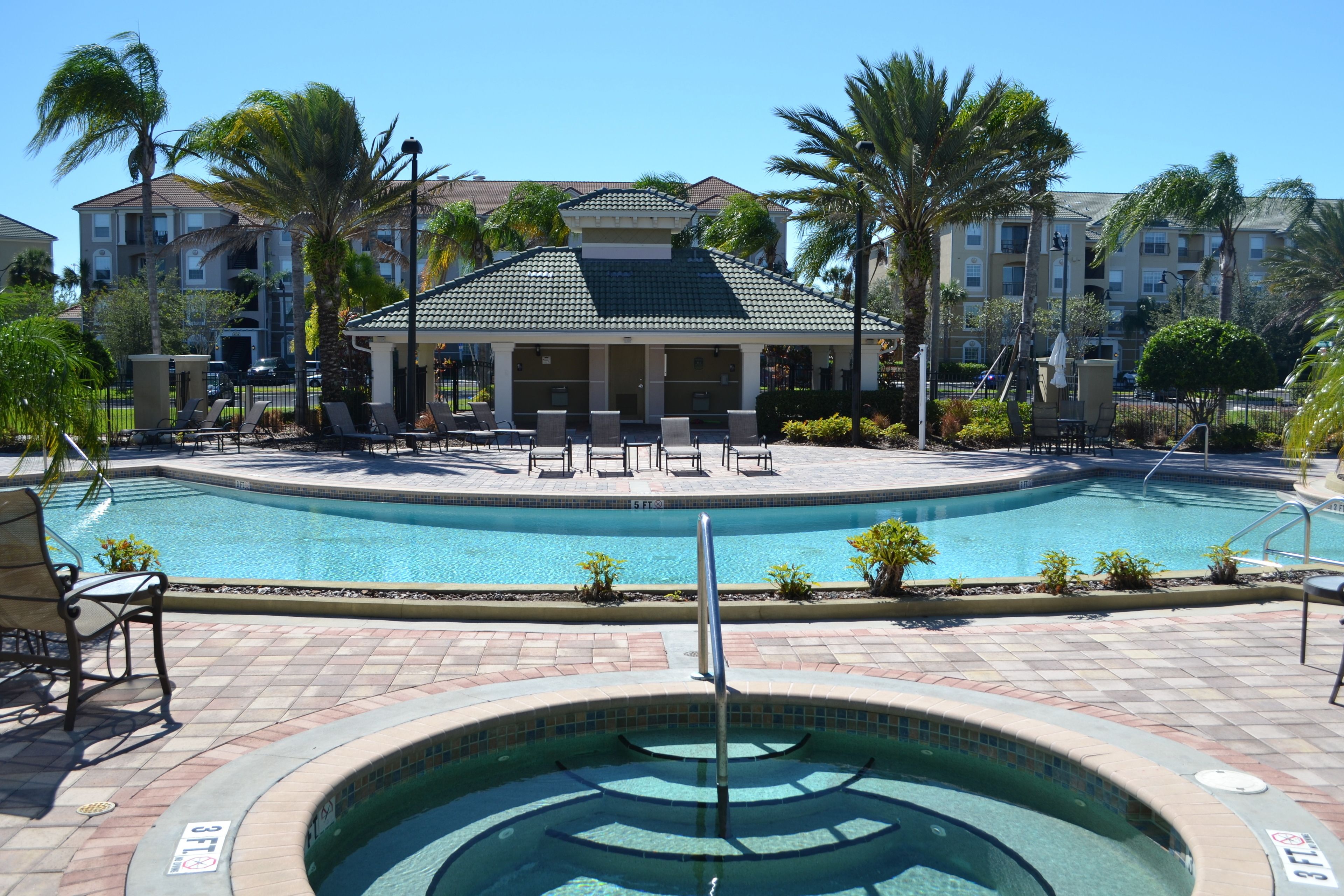 A pool area with lounge chairs and a hot tub, surrounded by palm trees and a building with a tiled roof.