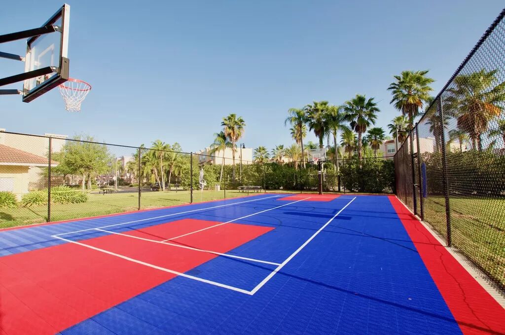Un campo da basket all'aperto con superficie blu e rossa, circondato da una recinzione e palme.