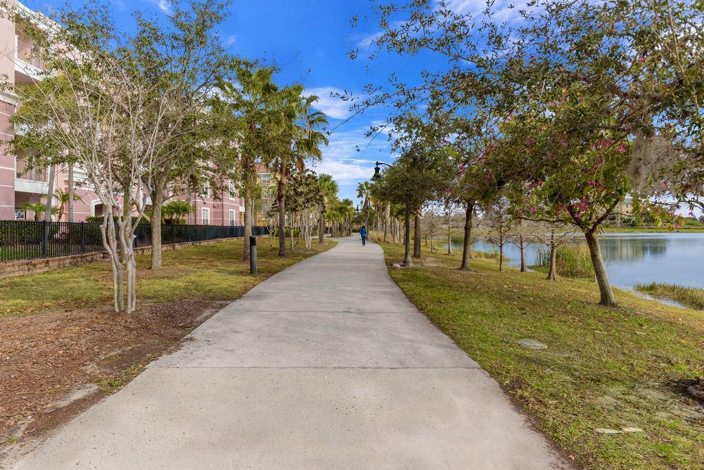 A concrete pathway flanked by trees and a body of water, leading to a building with balconies.