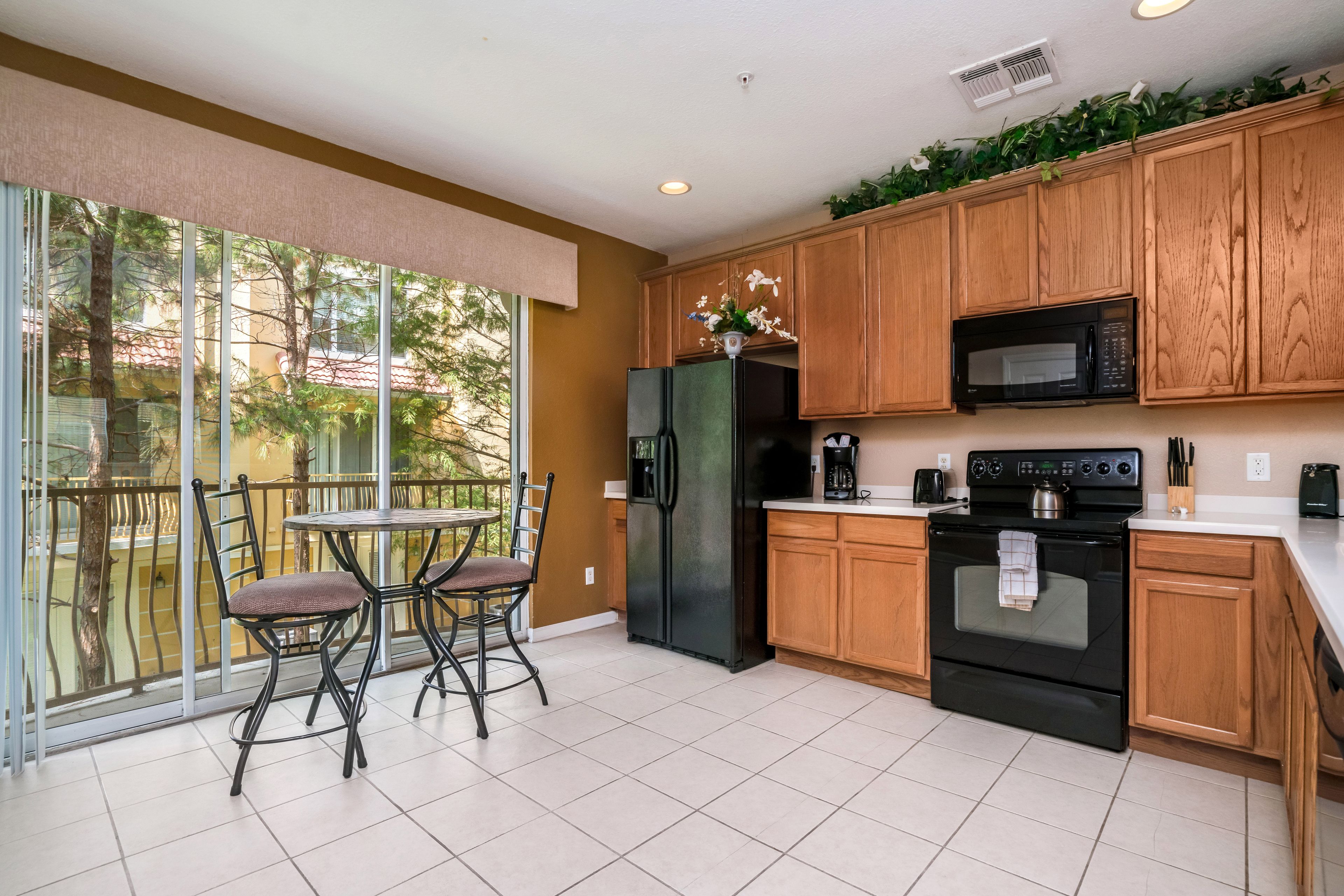 A kitchen with wooden cabinets, a black refrigerator, a microwave, and an oven. There is a dining table with chairs and a view of trees through the glass door.