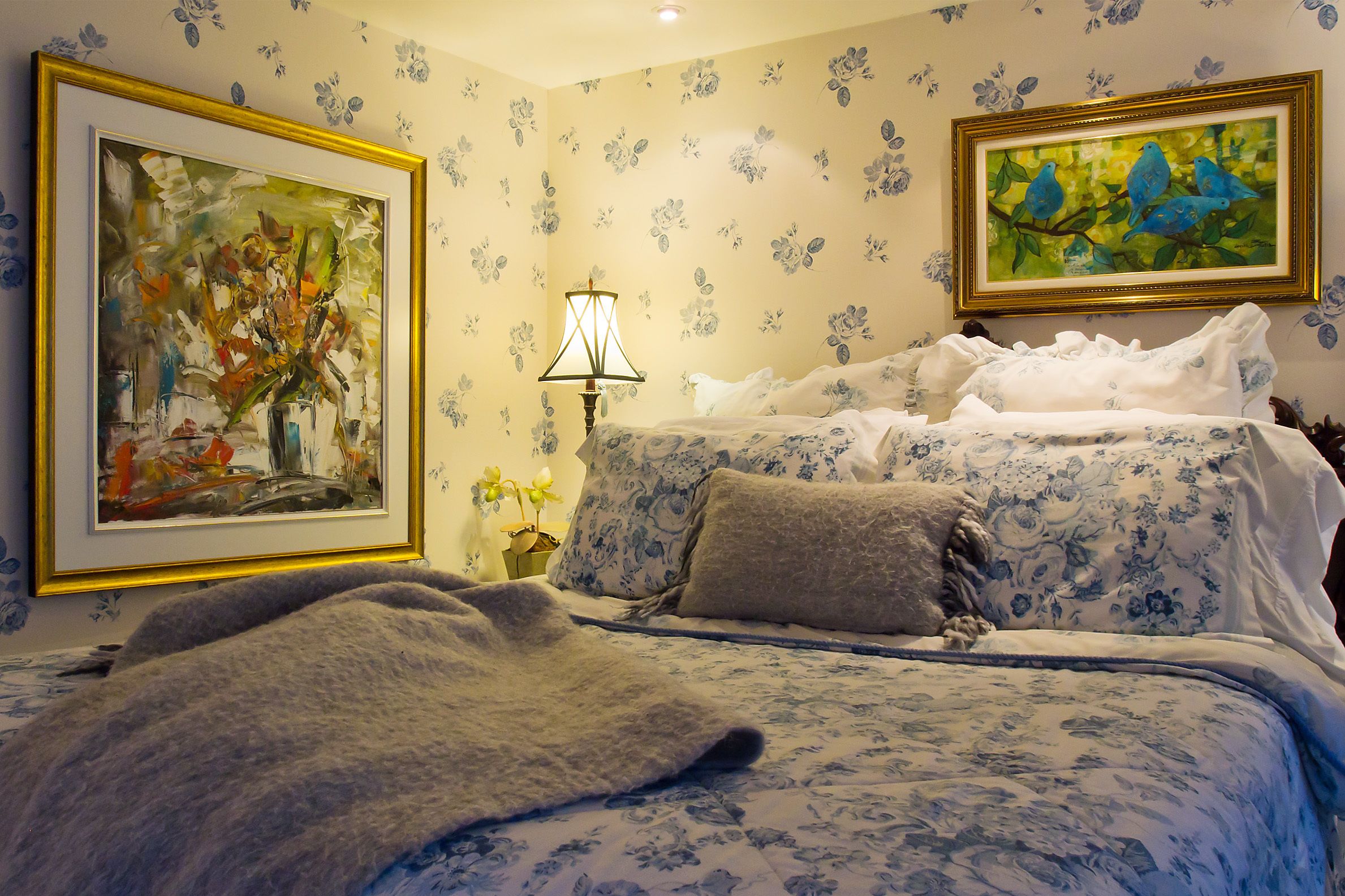 Un dormitorio con papel tapiz floral, una cama con cubrecama floral, dos almohadas, una mesita de noche con una lámpara y dos cuadros enmarcados.