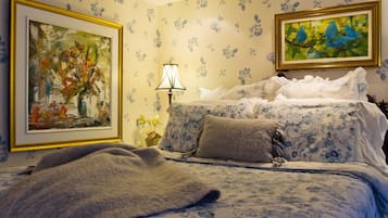 Un dormitorio con papel tapiz floral, una cama con cubrecama floral, dos almohadas, una mesita de noche con una lámpara y dos cuadros enmarcados.