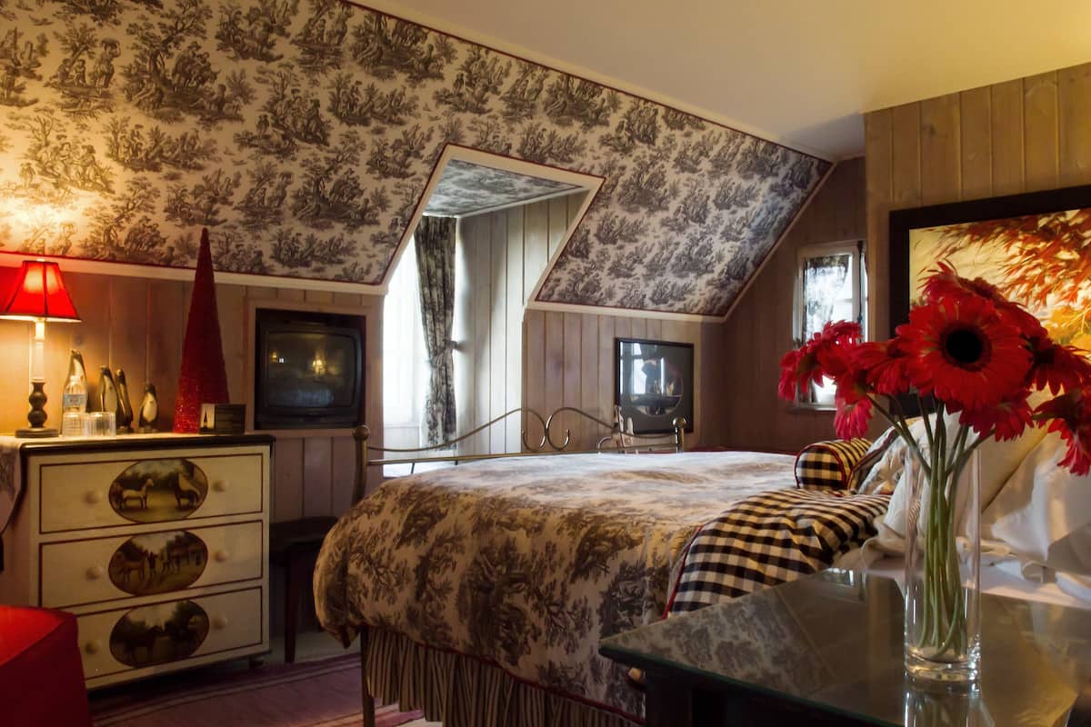 A bedroom with a sloped ceiling, a bed with a patterned bedspread, a dresser with decorative drawers, a red lamp, a television, and a vase with red flowers.