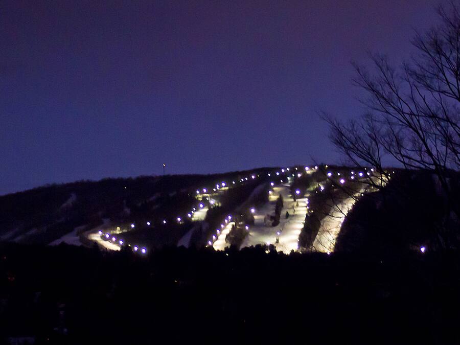 A nighttime view of a mountain with illuminated structures, possibly a resort or hotel, and a barren tree in the foreground.