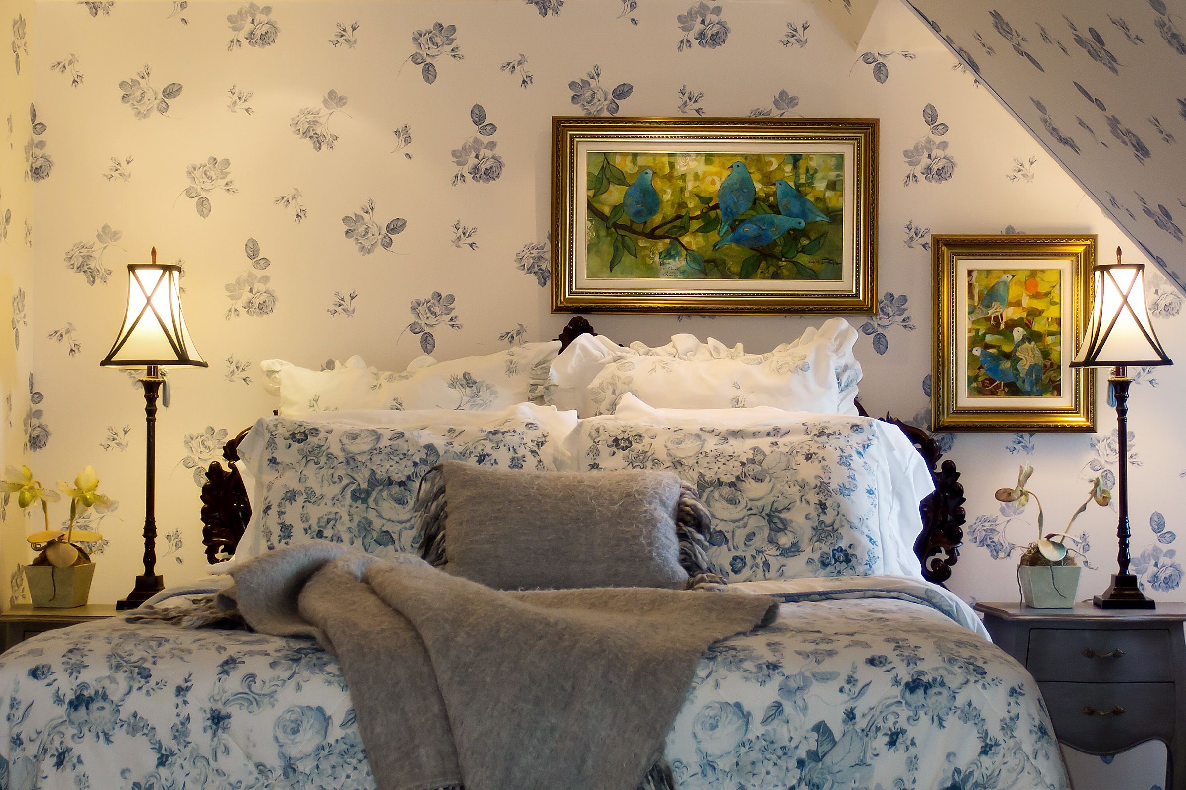 Une chambre à coucher avec un papier peint à motifs floraux, un lit recouvert d’une couverture grise, deux lampes de chevet et deux tableaux encadrés.