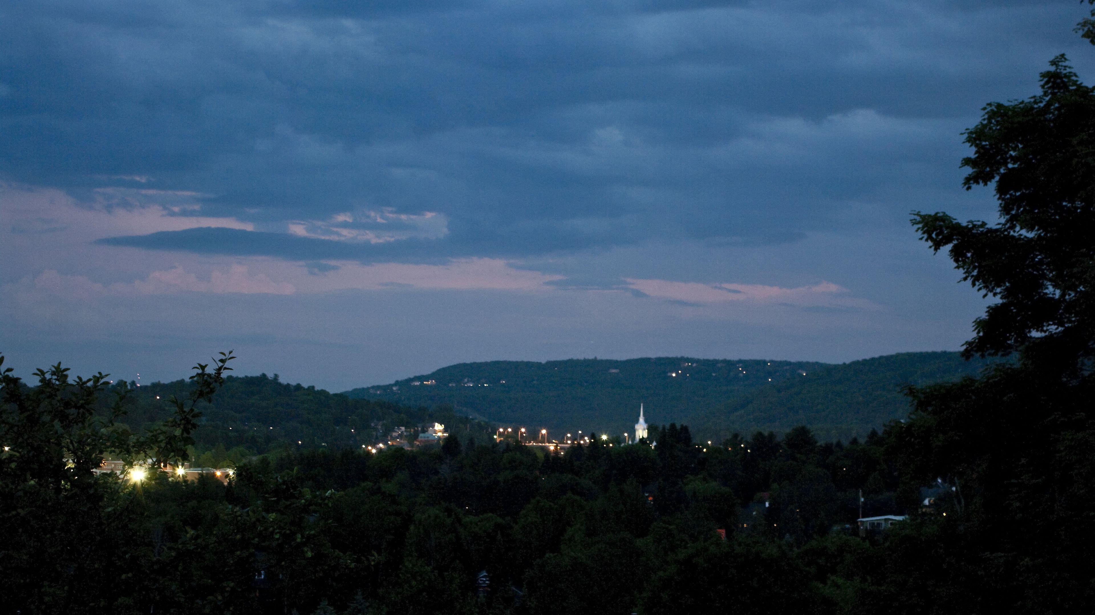 A nighttime view of a town with a prominent church spire, surrounded by hills and dense foliage.