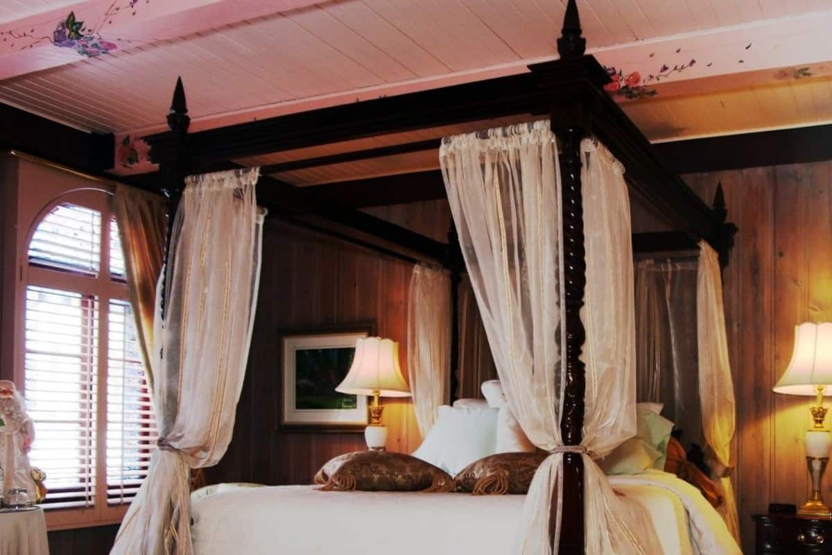 A four-poster canopy bed with white linens and a wooden headboard. There are two bedside lamps and a framed picture on the wall.