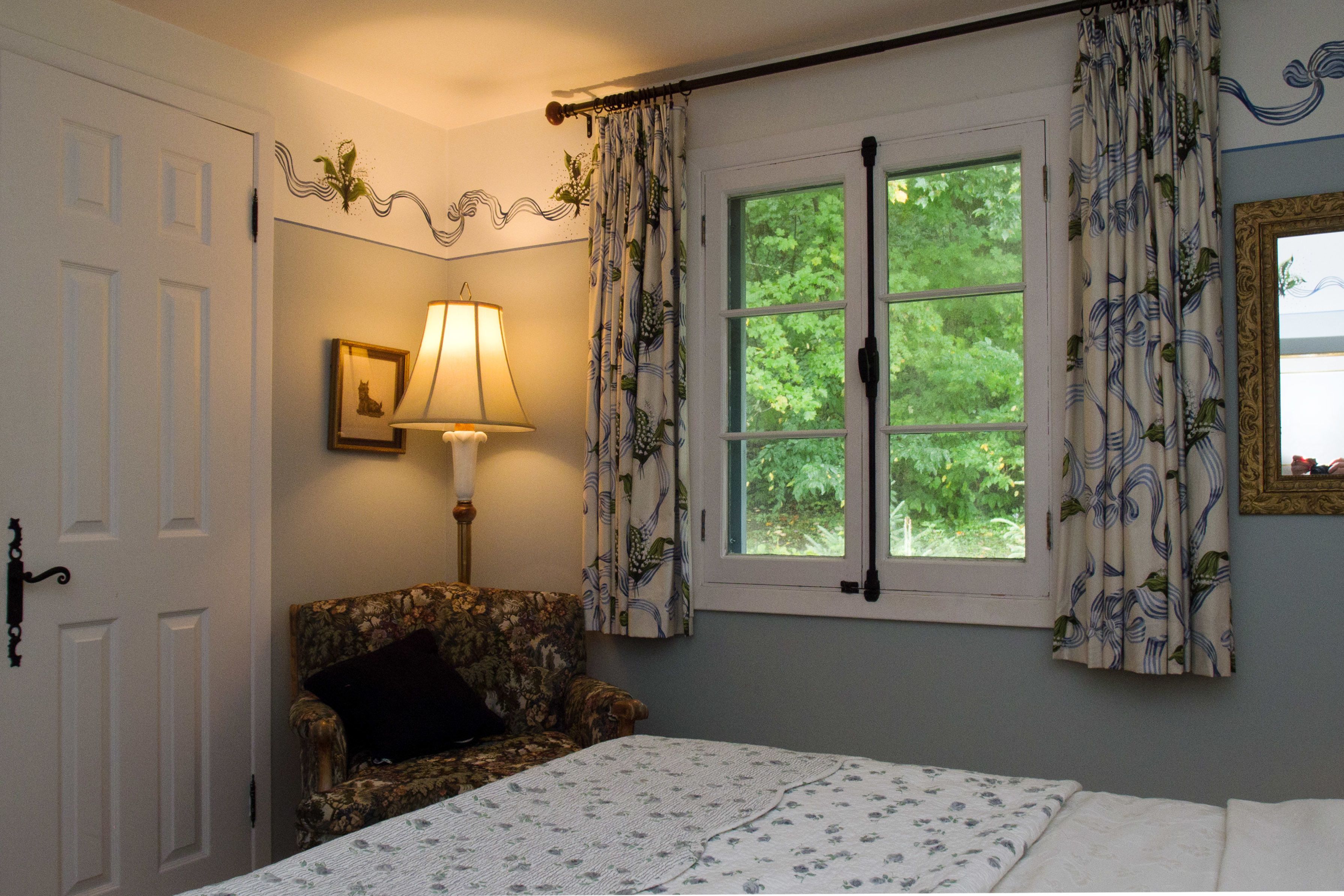 Un dormitorio con cama, una silla, una lámpara, una ventana con cortinas y una puerta.