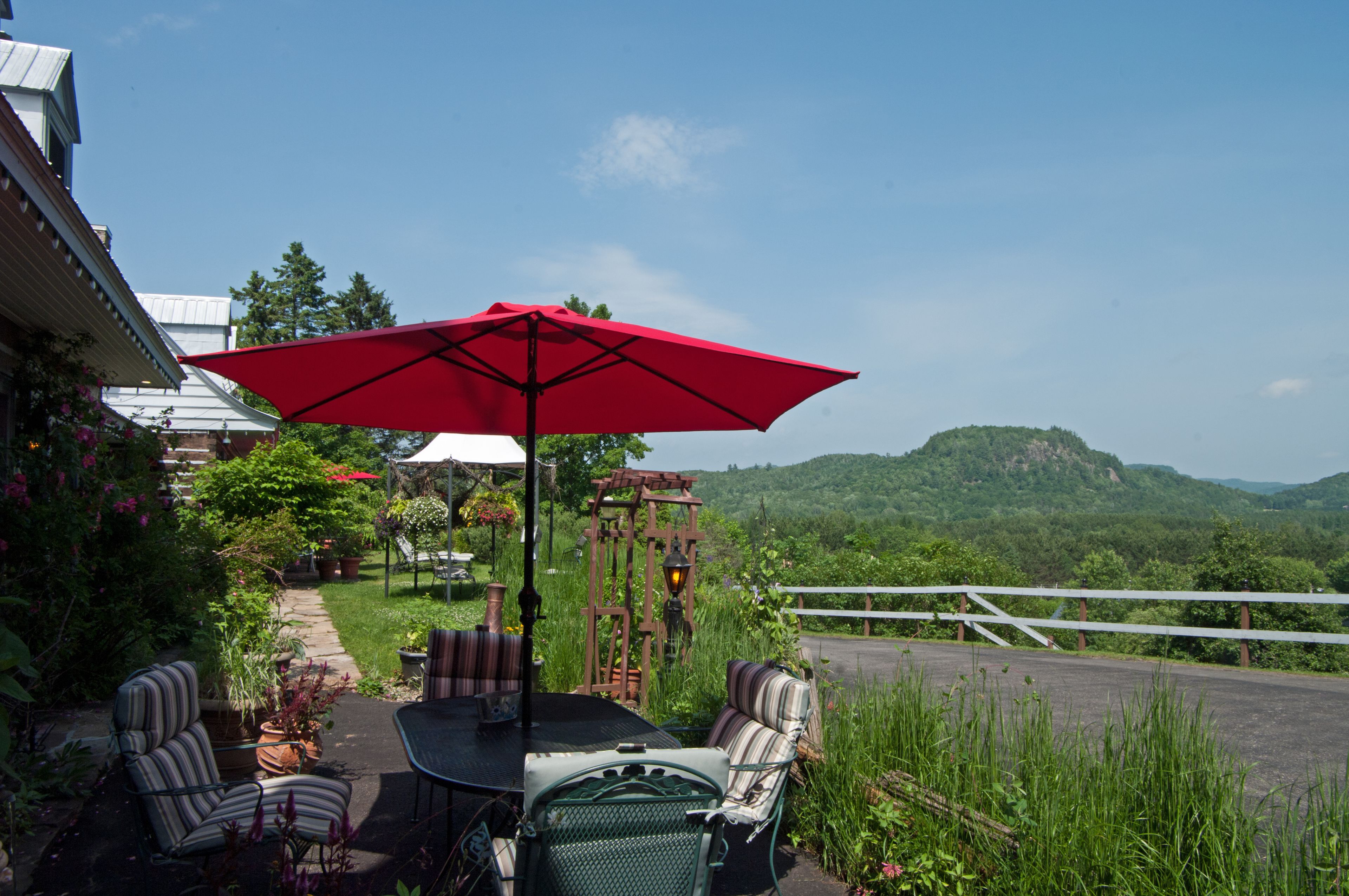 Un patio al aire libre con una mesa, sillas, un paraguas rojo y vistas a una montaña boscosa.