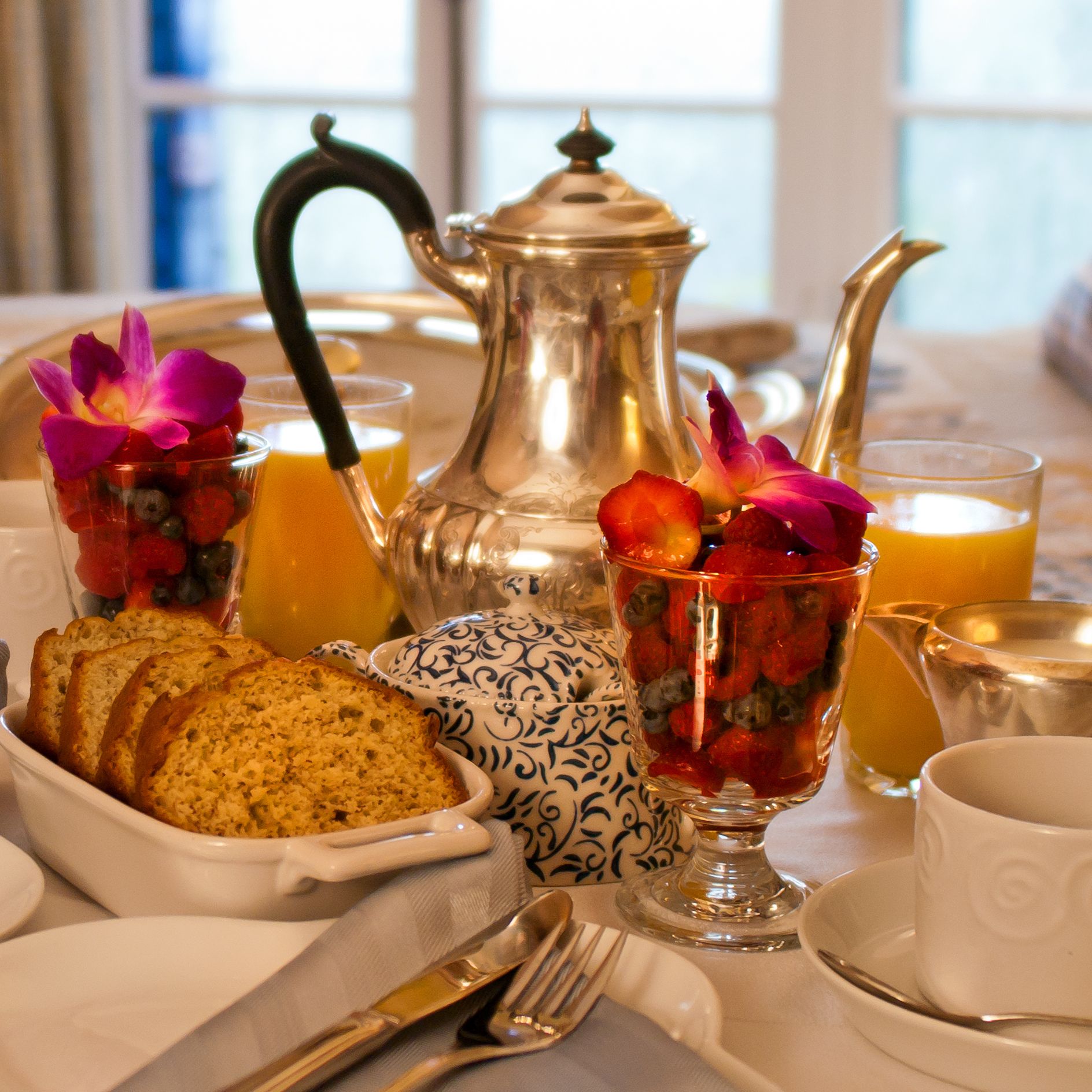 Una escena de desayuno con una tetera de plata, dos tazas de fruta, un bol de pan y una tetera con tapa.