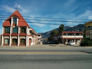 Un immeuble de deux étages, avec un toit rouge et un balcon, situé à l’angle d’une rue, avec une chaîne de montagnes en arrière-plan.