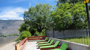 Une rangée de fauteuils de jardin verts, dotés d’un rembourrage confortable, longe un chemin de béton, avec une rivière et des montagnes en arrière-plan.