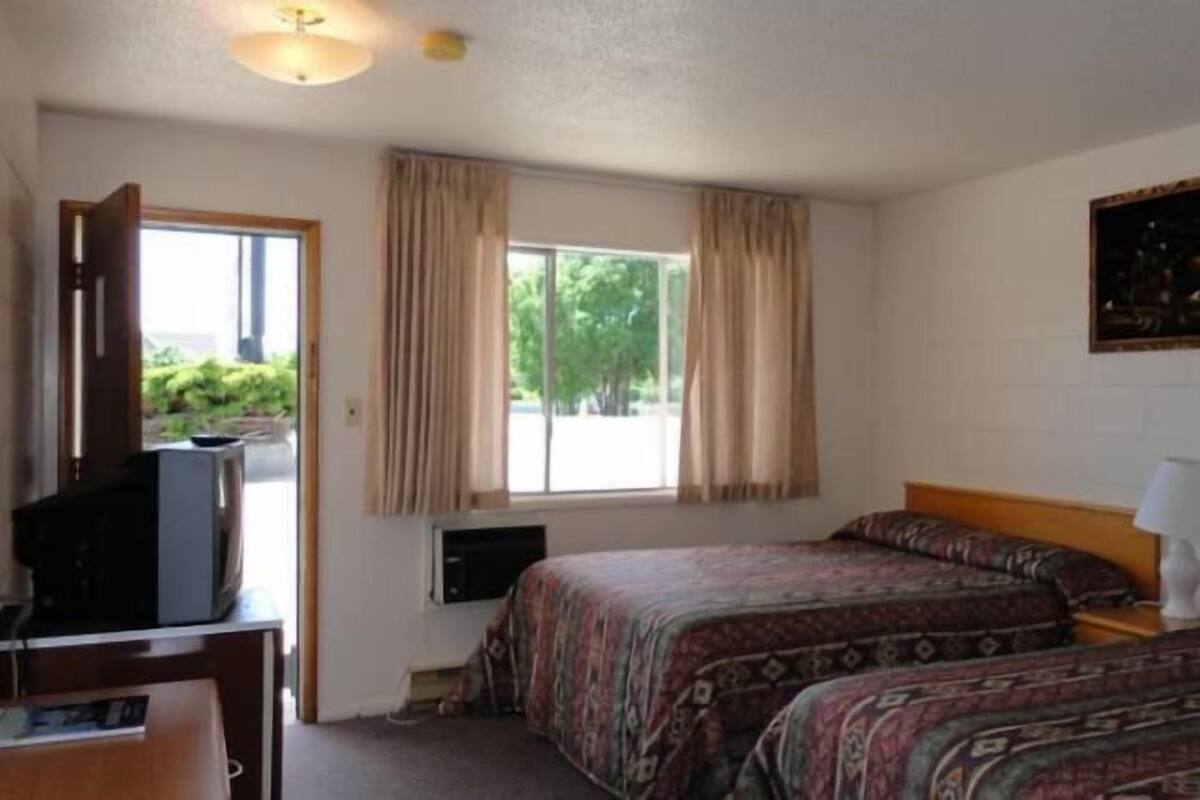 A hotel room with two beds, a desk with a television, a window with curtains, and a painting on the wall.