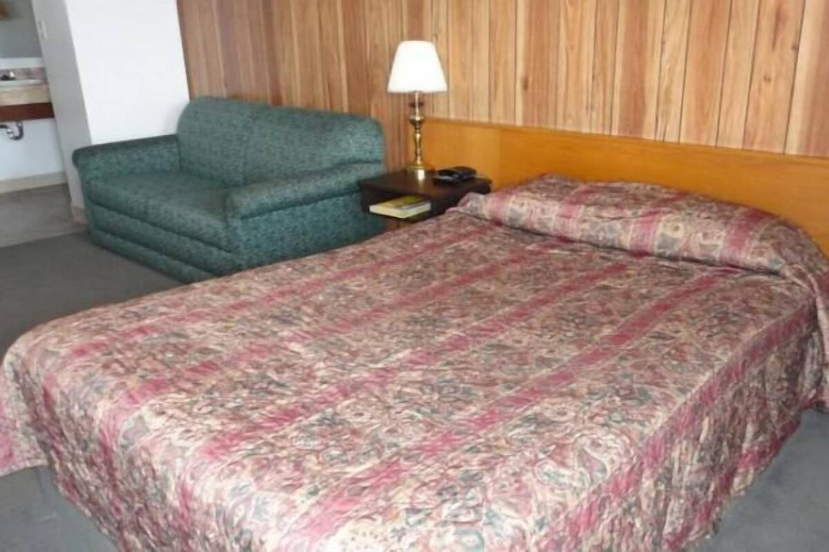 A hotel room with a bed, a green armchair, a nightstand with a lamp, and a wooden headboard.
