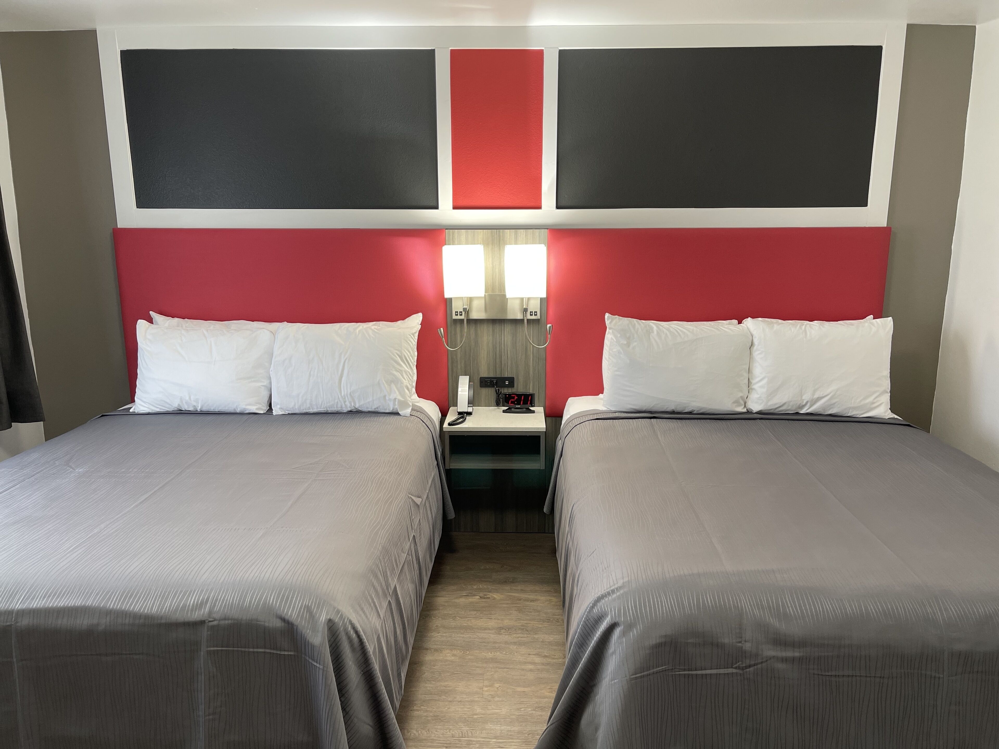 Habitación de hotel con dos camas, una mesita de noche con un teléfono y un reloj, y una pared de acento roja.