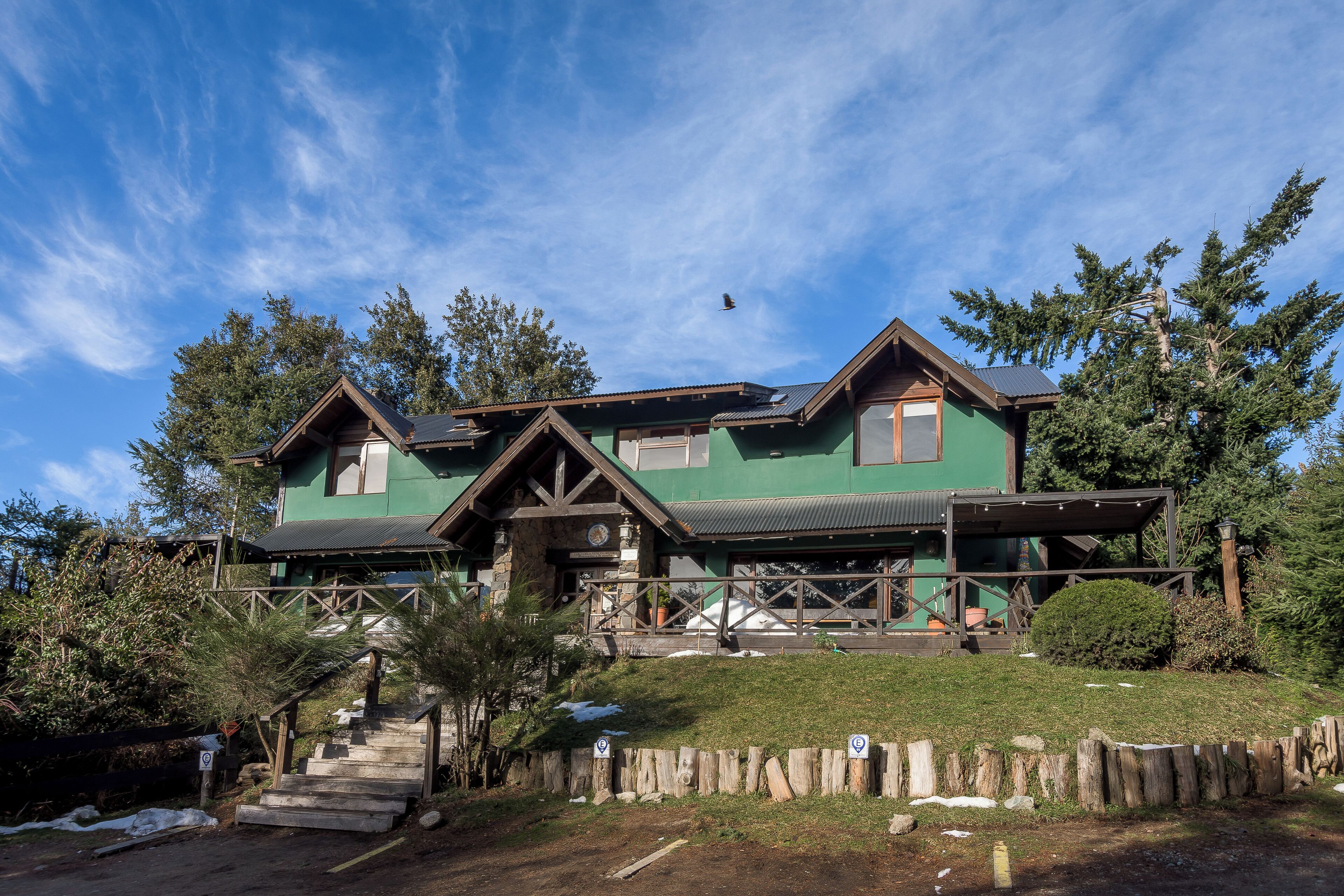 A two-story house with a green exterior and wooden accents, surrounded by trees and a wooden fence.