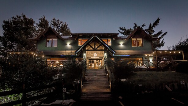 A two-story lodge with a central entrance, surrounded by trees and a wooden fence.
